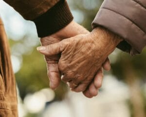 Two elderly people holding hands gently outdoors.
